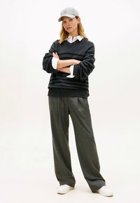 Oversized donkergrijze gestreepte trui, grijze pet, wit hemd met kraag, losse grijze broek, witte sneakers, staande houding.