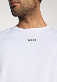 Λευκή αθλητική μπλούζα με υφή, με στρογγυλή λαιμόκοψη και μαύρο λογότυπο "BOSS" στο στήθος. Διακριτές κατακόρυφες ρίγες κατά μήκος των ώμων.