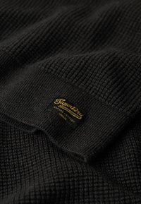 Svart stickad tyg med ett strukturerat mönster; har en vävd etikett i guld och svart som anger "Superdry" med platsinformation.