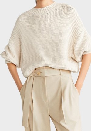 Femme portant un pull en maille crème ample avec des manches retroussées et un pantalon beige taille haute avec un détail bouton et ceinture.