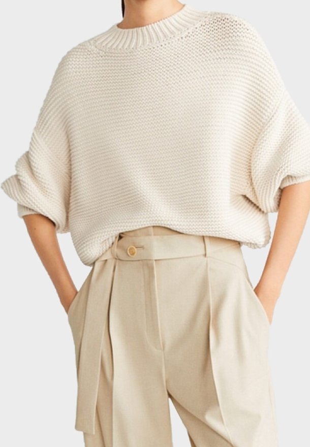 Femme portant un pull en maille crème ample avec des manches retroussées et un pantalon beige taille haute avec un détail bouton et ceinture.