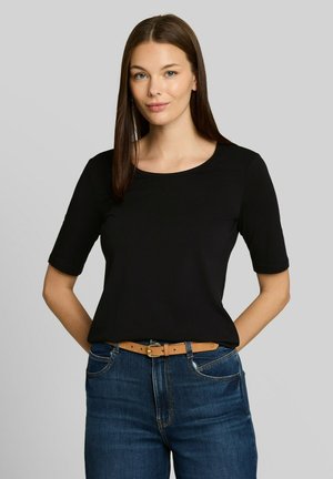 Junge Frau mit glattem braunem Haar, die ein schwarzes Kurzarmoberteil, blaue Jeans und einen hellbraunen Gürtel vor grauem Hintergrund trägt.