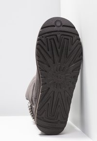 Botte en daim gris vue par en dessous, avec une semelle en caoutchouc texturée à motif en zigzag et une chaîne métallique décorative sur le côté.