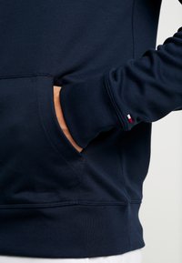 Granatowa bluza o gładkiej fakturze, z kieszenią na przodzie i ściągaczami przy rękawach, podkreślona małym detalem logo.