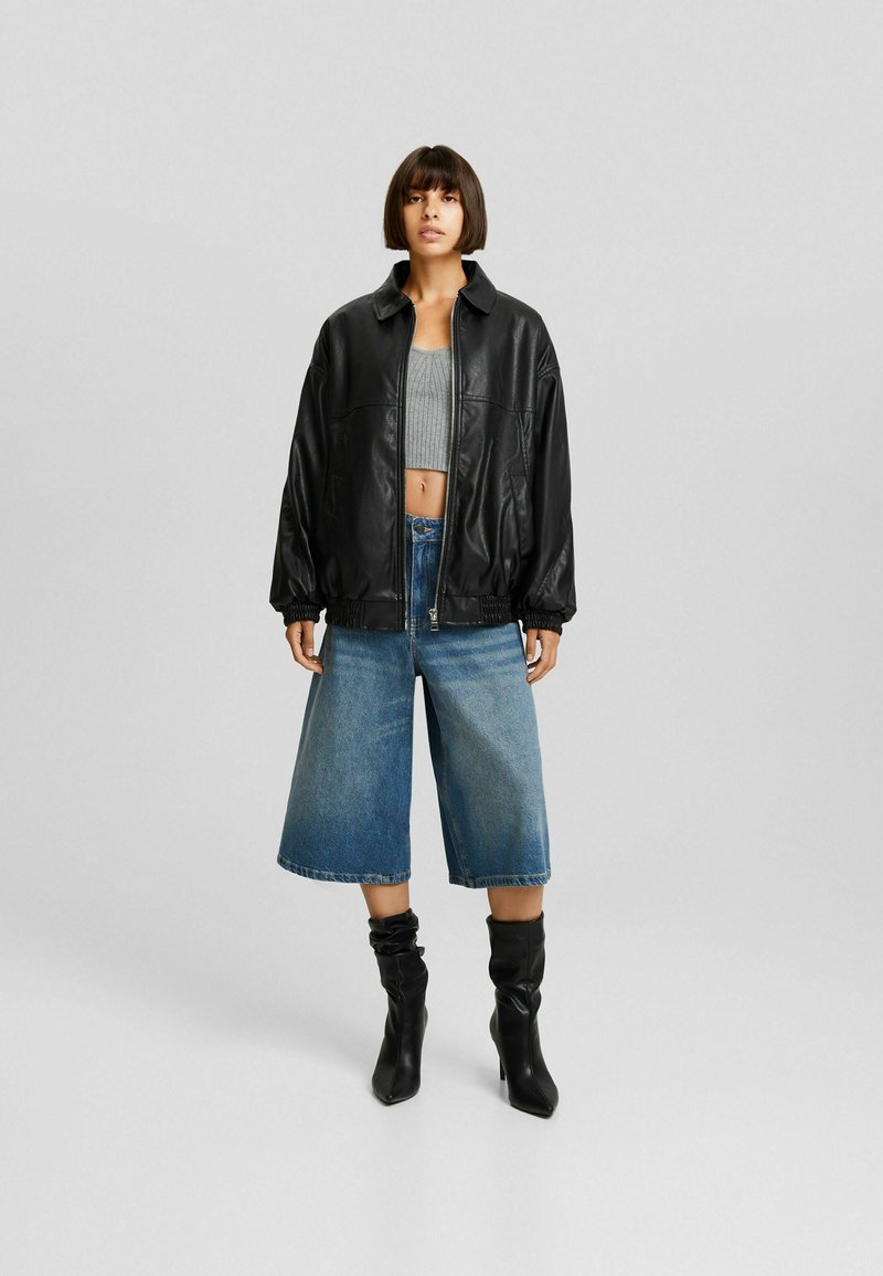 Mujer vistiendo una chaqueta de cuero negra, un top gris corto, unos pantalones cortos de mezclilla anchos hasta la rodilla y botas de tacón negras, de pie sobre un fondo gris.