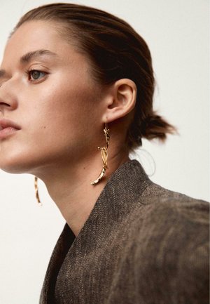 Woman with slicked-back hair wearing textured brown blazer and large abstract gold drop earrings, facing left against a plain background.