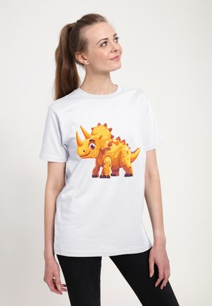 Kvinde står og smiler iført en hvid T-shirt med en tegneserieorange triceratops-dinosaur på forsiden.