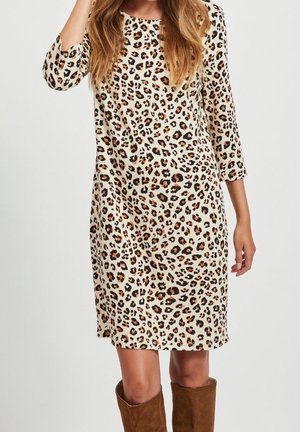 Kobieta w sukience o długości do kolan z długim rękawem w panterkę, połączona z brązowymi butami do połowy łydki, stojąca na jednolitym tle.