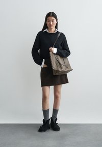 Modella in un maglione nero a maglia con polsini a coste, una camicia bianca sotto, una gonna marrone, calze grigie fino al ginocchio e stivali neri, mentre tiene una borsa con motivo.