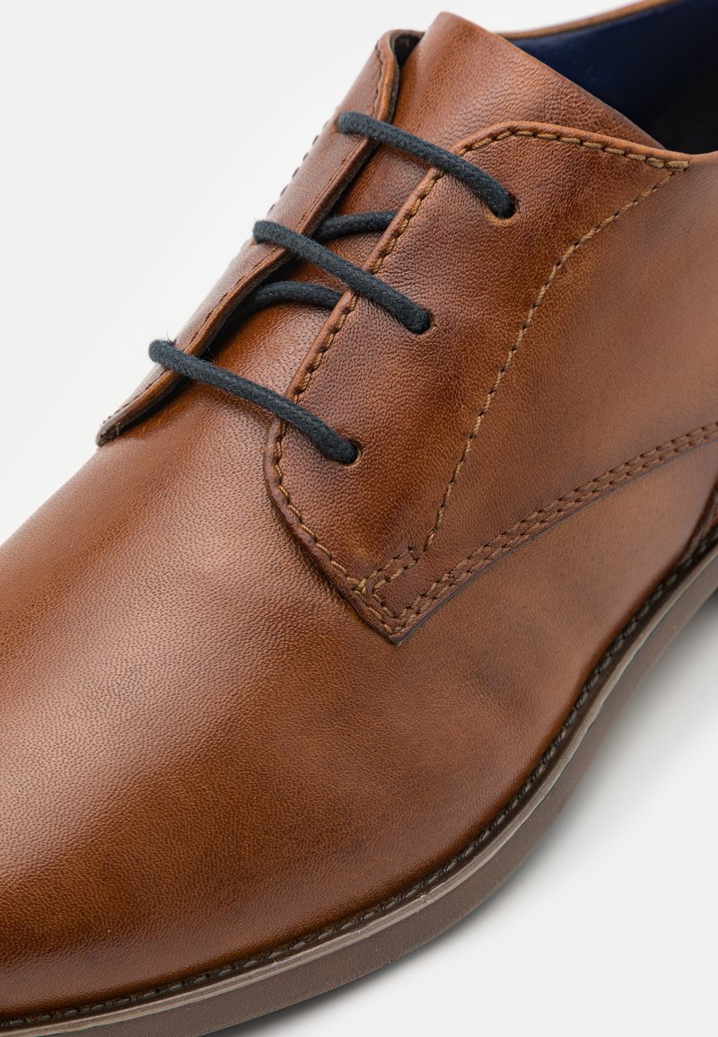 Braunes Leder-Schuh mit glatter Oberfläche, ausgestattet mit schwarzen Schnürsenkeln und kontrastierenden Nähten. Rundliche Zehenform und flache Gummisohle.