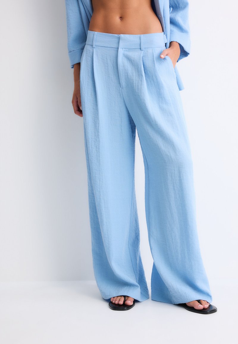 Personne portant un pantalon ample bleu clair, une veste ouverte assortie et des sandales noires, debout devant un fond clair uni.