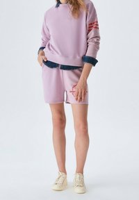Person som bär en lila sweatshirt och shorts-set med röd text, lager på ett denimskjorta, tillsammans med krämfärgade sneakers, stående framför en vit bakgrund.