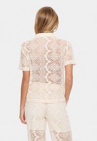 Beige spetsblus med korta ärmar, med intrikata mönster och en genomskinlig baksidesdesign som visar en texturerad, virk-liknande utseende.