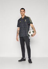 Fekete mintás futballmez arany díszítéssel, fekete nadrággal, amelyen fehér oldalsávok találhatók, és fekete sportcipő. Futballlabdát tart a kezében.