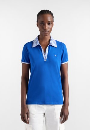 Vrouw die een blauw poloshirt met korte mouwen draagt, met gestreepte kraag en manchetten, staand tegen een effen witte achtergrond.