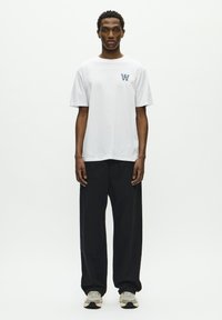 White cotton t-shirt with a round neckline and short sleeves, featuring a small navy logo. Paired with black loose-fitting trousers and gray sneakers.