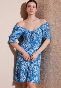 Rochie cu umerii descoperiți, în model floral albastru și alb, dintr-un material texturat, cu mâneci puff și un detaliu de strângere cu fundă la guler.