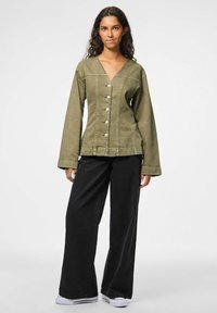 Giovane donna con lunghi capelli scuri, che indossa una giacca verde oliva con bottoni, pantaloni neri larghi e sneakers bianche su uno sfondo neutro.