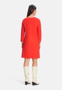 Rotes knielanges Kleid mit Dreiviertelärmeln und einem Reißverschluss im Rücken, kombiniert mit weißen kniehohen Stiefeln mit Blockabsatz.