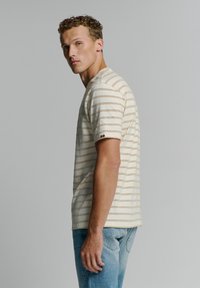 Beige gestreiftes T-Shirt mit kurzen Ärmeln und rundem Halsausschnitt, ausgestattet mit horizontalen Linien und einem kleinen Logo-Akzent am Ärmel. Trägt dazu hellblaue Jeans.