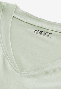 Helles grün, T-Shirt mit V-Ausschnitt und Cap-Ärmeln aus weicher Baumwolle. Besitzt eine glatte Textur und sichtbare Nähte entlang des Halsauschnitts. Markenlabel "NEXT" vorhanden.