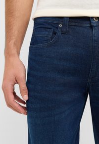 Vaqueros de denim azul oscuro con un ajuste clásico. Las características incluyen un bolsillo frontal, detalles de costura en la parte superior y una textura de tela suave.