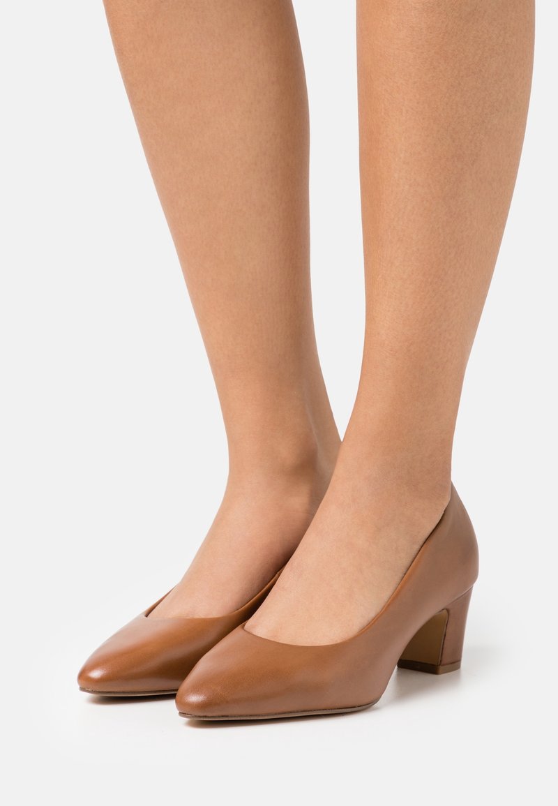 Jambes et pieds chaussés de escarpins en cuir marron à talons bas, bout fermé, sur fond blanc.