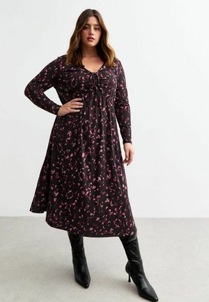 Robe noire à manches longues avec un motif abstrait rose, décolleté en V, détails de lien réglables et jupe évasée, associée à des bottes noires hautes jusqu'aux genoux.