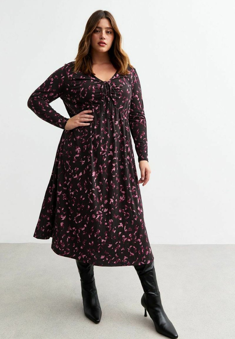 Vestido negro de manga larga con un patrón abstracto en rosa, escote en V, detalles de lazo ajustables y falda acampanada, combinado con botas negras hasta la rodilla.