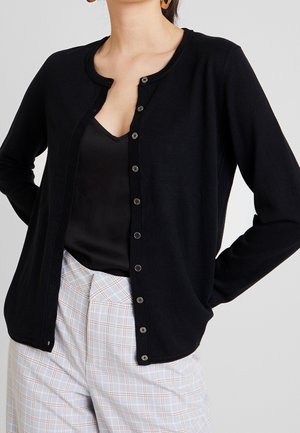 Vrouw draagt een zwarte cardigan met knopen over een zwart shirt, gecombineerd met licht geblokte broek, handen op de heupen.