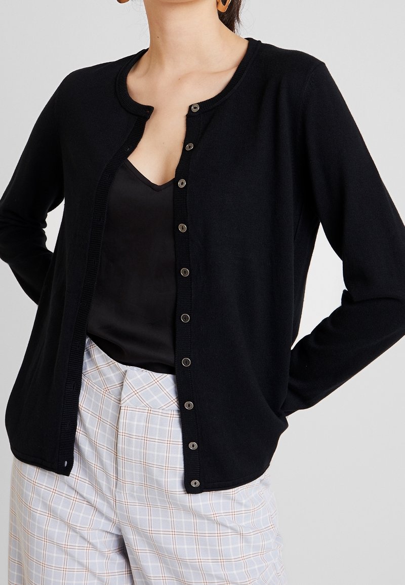 Femme portant un cardigan noir boutonné sur un haut noir, associée à un pantalon à carreaux clair, les mains posées sur les hanches.