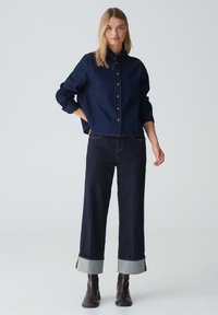 Giacca in denim scuro con chiusura a bottoni, maniche lunghe e colletto. Abbinata a jeans larghi scuri, con orli risvoltati in denim più chiaro.