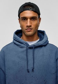 Blauwe hoodie met een gestructureerde stof, voorzien van een relaxte pasvorm, koordjes en versleten details. Draagt met een zwarte pet.