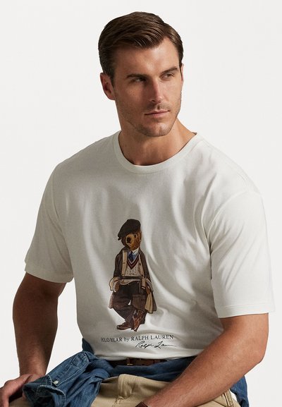 Muž v bielom tričku s ilustrovaným medveďom oblečeným vo vintage oblečení a nápisom "Polo Bear by Ralph Lauren" sedí s modrou bundou na lone.