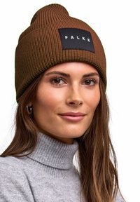 Cappello a maglia marrone con texture a coste e punta superiore, con una toppa nera con la scritta "FALKE". Indossato con un maglione dolcevita grigio chiaro.