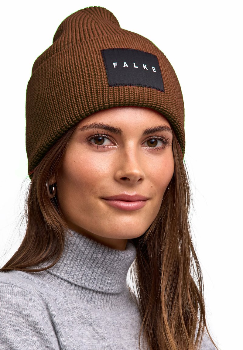 Cappello a maglia marrone con texture a coste e punta superiore, con una toppa nera con la scritta "FALKE". Indossato con un maglione dolcevita grigio chiaro.