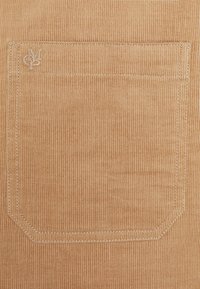 Tessuto in velluto a coste color tan con una tasca rettangolare dotata di cuciture precise e un piccolo logo ricamato nell'angolo superiore.