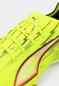 Jasnożółty but piłkarski z fakturowaną powierzchnią, czarnym wzorem błyskawicy i czerwonym akcentem wzdłuż boku oraz pasującymi sznurowadłami.