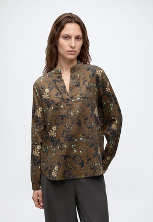 Femme portant un chemisier à manches longues marron à motifs floraux avec une patte de boutonnage partielle sur le devant et un pantalon noir, debout devant un fond uni.