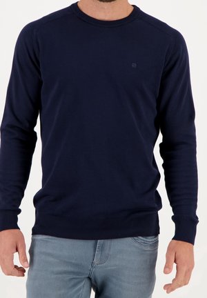 Homme portant un pull simple à manches longues bleu marine foncé et un jean bleu clair, debout devant un fond blanc uni.