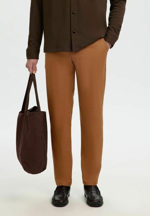 Personne portant un pantalon marron et une chemise marron foncé, tenant un sac fourre-tout marron foncé dans une main, avec des chaussures noires visibles.