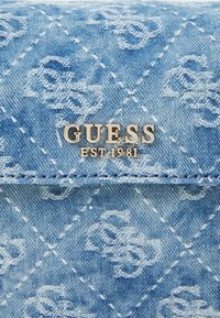 Džínová peňaženka vo svetlomodrej farbe s embosovaným logom a prešívaným vzorom. Strieborné kovanie výrazne zobrazuje "GUESS EST 1981".