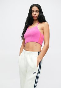 Mulher com cabelo preto comprido veste um top halter rosa curto e calças de treino brancas da Adidas com riscas em azul-marinho, posando com as mãos nos bolsos.