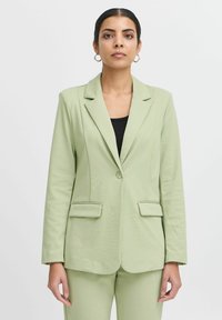 Světle zelený blazer z texturovaného materiálu, s jedním knoflíkem, dvěma předními kapsami, přiléhavým střihem, klopovým límečkem a dlouhými rukávy.