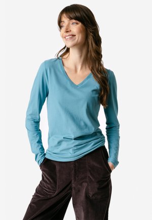 Frau mit langen braunen Haaren, die ein blaues, langärmeliges V-Neck-Shirt und dunkle Hosen trägt, lächelt und schaut zur Seite, die Hände in den Taschen.