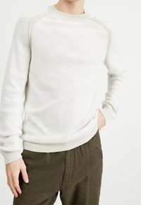 Jasnobeżowy sweter z dzianiny o fakturowanej tkaninie, z okrągłym dekoltem i prążkowanymi mankietami. Połączony z brązowymi spodniami w kratę.