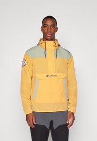 Letvægts gul og grøn vindjakke med en frontlomme, justerbar hætte og Columbia-logo på brystet. Strukturstof, afslappet design.