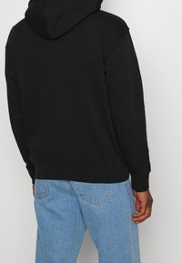 Černá bavlněná mikina s kapucí na šňůrku, žebrovanými manžetami a lemem, v kombinaci se světlými modrými džínami. Hladká textura s minimálními detaily.