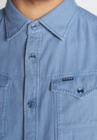 Ljusblå jeansskjorta med spetsig krage, framfickor och mörka knappdetaljer. Har en vävd textur och en märkesetikett.