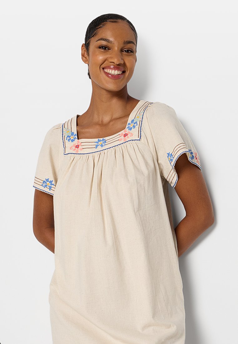 Beige linnen jurk met korte mouwen, voorzien van kleurrijke bloemenborduursels en een vierkante halslijn. De stof heeft een zachte, gestructureerde uitstraling.